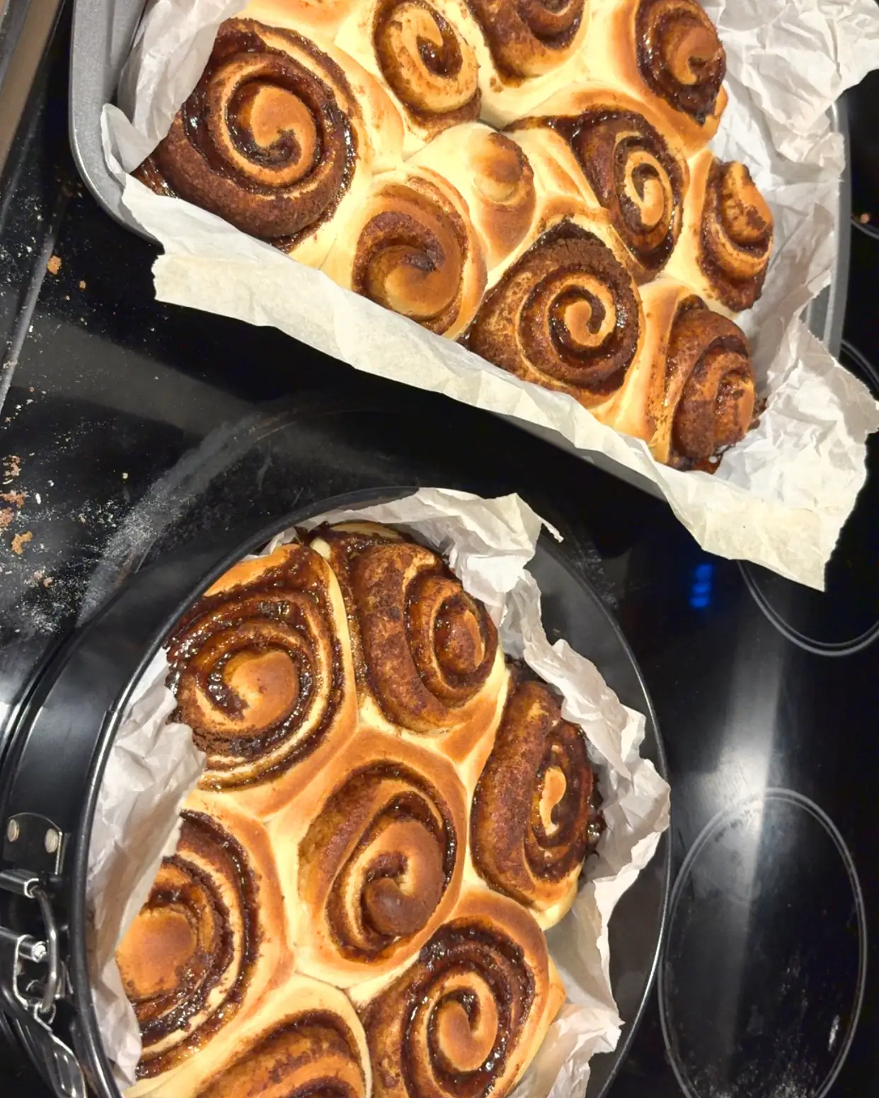 Two pans of baked cinnamon rolls on a black stovetop, a rectangular tin above and a round springform pan below, both lined with parchment.